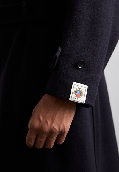 Námořnický vlněný kabát s knoflíkovým manžetem, s bílou cedulkou s barevným znakem a textem "BALLY". Hladká textura a propracovaný střih.