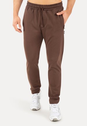 Mand iført brune elastiske joggingbukser med snørre og hvide atletikskøjter, hånd i lommen, stående på hvid baggrund.