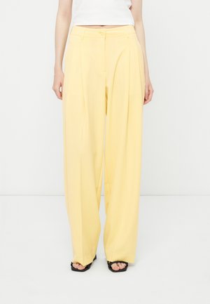 Person wearing high-waisted, loose-fitting pale yellow pants with front pleats and black open-toe sandals, standing against a plain background.