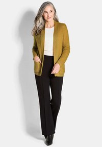 Senfgelber Cardigan mit strukturiertem Strick, offener Front und zwei Fronttaschen, kombiniert mit schwarzen Hosen. Leichter Stoff und entspannte Passform.