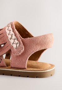 Sandalo in suede rosa con dettagli intrecciati metallici, design aperto e soletta imbottita. La suola è marrone testurizzata con un piccolo tacco.