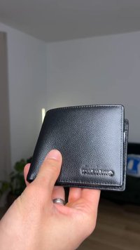 Zwarte leren portemonnee met een textuur oppervlak, bi-fold ontwerp en geëmbossed merk op de voorkant. Gehouden in de hand tegen een neutrale achtergrond.