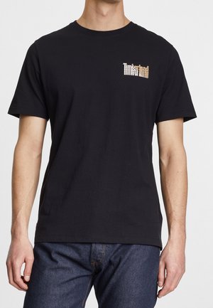 T-shirt noir à manches courtes avec col rond et petit logo Timberland sur la poitrine gauche, porté avec un jean bleu foncé.