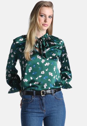 Vrouw met lang blond haar, gekleed in een groene satijnen gebloemde blouse met een striksluiting bij de hals en blauwe spijkerbroek met een zwarte riem tegen een egale achtergrond.
