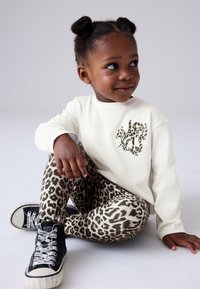 Camiseta blanca de manga larga con un logotipo de estampado de leopardo, combinada con leggings color beige con un patrón de leopardo en negro y marrón, y zapatillas negras de caña alta.