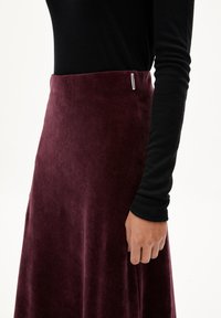 Personne portant un haut noir à manches longues et une jupe en velours noir violet taille haute avec une petite étiquette blanche sur la ceinture.