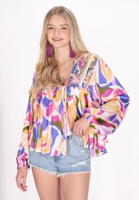 Multifarvet bluse med en løs, svævende design og detaljeret broderi, ballonærmer og en bindelukning ved halsen. Sammen med denimshorts.