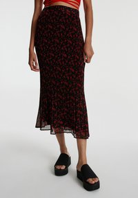 Femme portant une jupe midi noire à motif floral rouge, un crop top rouge à bretelles et des sandales plateformes noires à bout ouvert, debout sur fond blanc.