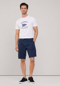 T-shirt bianca in cotone con stampa grafica blu, abbinata a pantaloni corti navy in tessuto leggero e sneakers nere. Design semplice.