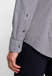 Černobílá kostkovaná košile s manžetami na knoflíky, s kontrastními knoflíky a hladkou texturou tkaniny. Manžeta přesahuje zápěstí.