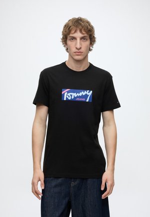 Giovane uomo con capelli biondi ondulati che indossa una t-shirt nera Tommy Jeans e jeans blu scuro, in piedi davanti a uno sfondo bianco semplice.