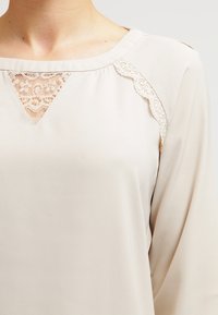 Blouse beige avec un col rond, délicate bordure en dentelle dans une découpe triangulaire, et tissu doux et lisse avec une coupe décontractée.