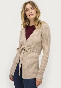 Cardigan a costine beige chiaro con cintura, caratterizzato da maniche lunghe e scollatura a V, indossato sopra una maglia rosso scuro e jeans blu.