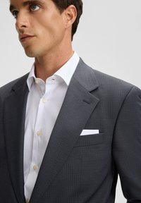 Młody mężczyzna w ciemnoszarym żakiecie na białej koszuli, z białą poszetką w kieszeni piersiowej żakietu.