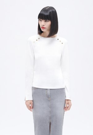 Top blanco de manga larga con escote redondo, adornado con clavos dorados en los hombros, combinado con una falda de mezclilla gris. Textura suave.