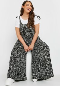 Svart blommig jumpsuit med vida ben och knytband, över en vit t-shirt. Har vita blommor mot en svart bakgrund.