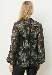 Blusa nera trasparente con motivo floreale metallico, maniche lunghe a sbuffo e chiusura con bottone al collo. Tessuto morbido, design fluido.