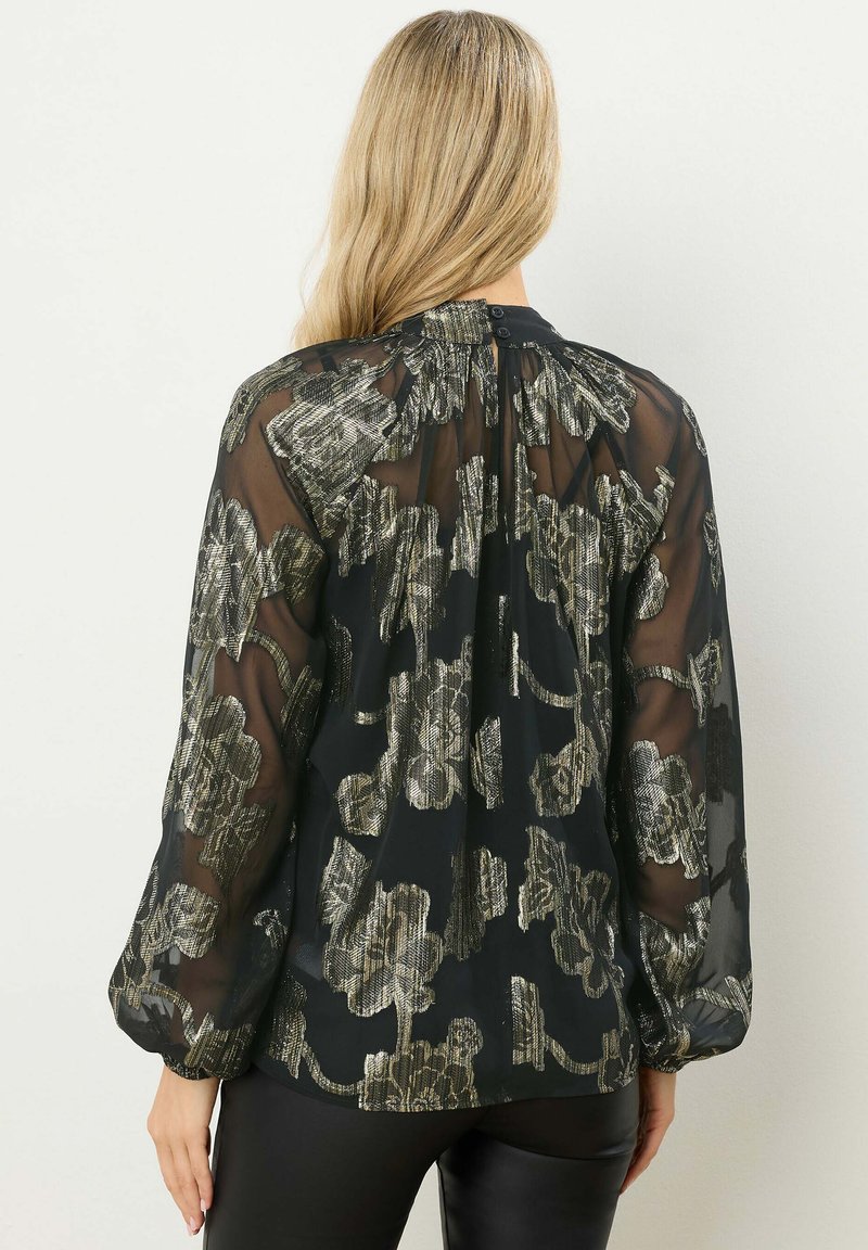 Blusa nera trasparente con motivo floreale metallico, maniche lunghe a sbuffo e chiusura con bottone al collo. Tessuto morbido, design fluido.