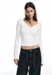 Witte nauwsluitende top met lange mouwen en een V-halsaccent met kanten bies, gecombineerd met donkerblauwe, hoog-taillierte spijkerbroek.