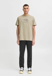 Khaki cotton t-shirt with short sleeves, featuring a printed design in black. Paired with black sweatpants and white sneakers.