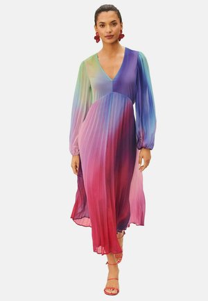 Vrouw draagt een lange, geplooide, ombré jurk met lange mouwen in tinten groen, paars en roze, gecombineerd met rode sandalen met bandjes en oorbellen.