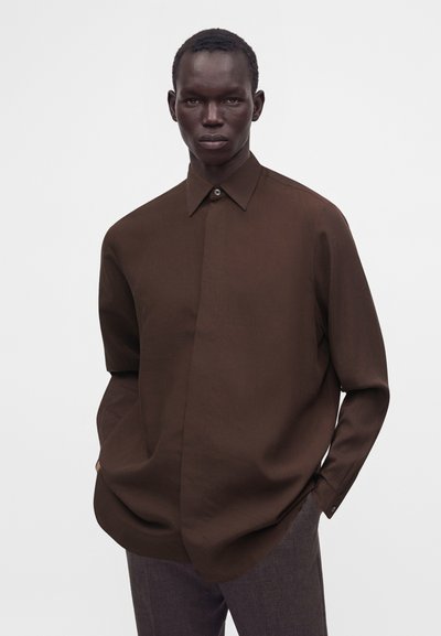 Homme à la peau foncée portant une chemise ample marron foncé boutonnée et un pantalon gris, debout les mains dans les poches devant un fond uni.