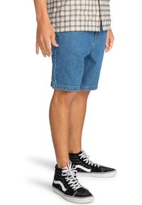 Man in blauwe denim shorts, zwarte hoge sneakers met witte veters, en een beige geruite shirt, staand op een witte achtergrond.