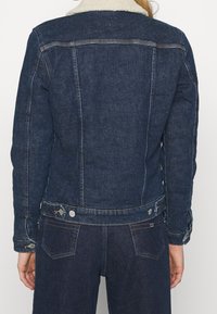 Person wearing dark blue denim jacket with white shearling collar and matching dark blue denim jeans, shown from the back.