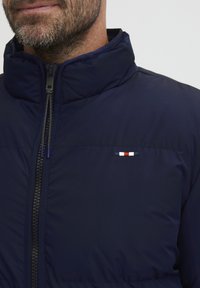 Navy Daunenjacke mit hohem Kragen, Frontreißverschluss und einem kleinen dreifarbigen Logo-Akzent an der Schulter. Glattes, wasserabweisendes Material.