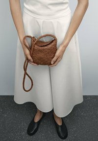 Woven brown leather bag with a curved top, slender strap, and intricate pattern. Held by a person in a white skirt, against a neutral background.