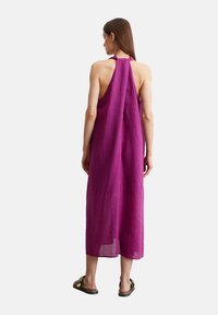 Robe sans manches en lin violet, longueur genou, coupe ample, avec un dos ouvert et des coutures minimales. Portée avec des tongs noires et un talon plat.