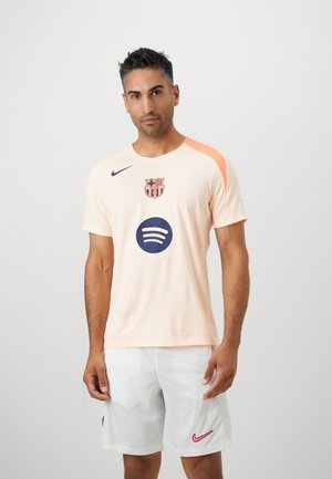 Man in een licht perzikkleurig FC Barcelona voetbalshirt met Spotify- en Nike-logo's, gecombineerd met witte Nike-shorts, staand tegen een effen achtergrond.
