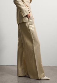 Pantaloni larghi in metallo dorato con una texture liscia, dotati di tasche frontali e spacco laterale, abbinati a ballerine a punta color crema.
