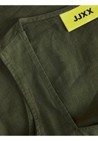 Prenda de color verde oliva con una tela texturizada y tejida. Presenta un escote limpio y una etiqueta amarilla con la inscripción "JJXX" cerca de la costura.