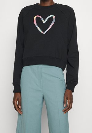 Person trägt einen schwarzen Sweatshirt mit langen Ärmeln und mehrfarbigem Herzdesign sowie hoch taillierte hellblaue, weit geschnittene Hosen.