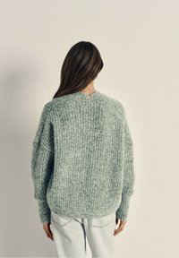 Overmaat, textuurtrui in lichtblauw-groene brei, met een V-hals en geribbelde manchetten, gedragen met lichte denim jeans.