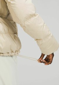 Main à la peau foncée ajustant le cordon de serrage de l'ourlet d'une doudoune beige sur un fond gris clair uni.