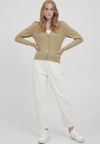 Cardigan beige côtelé à manches longues, avec un devant à boutons et une taille ajustée, associé à un pantalon blanc ample et des baskets blanches.