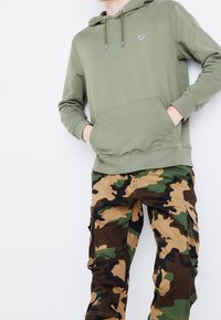 Persoon in een olijfgroene hoodie met een voorzak en camouflage cargo-broeken, staande tegen een witte achtergrond.