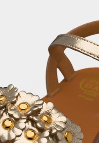 Εγγύς πλάνο ενός χρυσού σανδαλιού με μεταλλικά διακοσμητικά λουλουδιών και έναν μονό ιμάντα πάνω σε ταμπά πάτο.