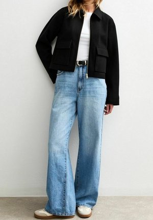 Vrouw draagt een zwarte cropped jas, wit topje, lichtblauwe hoge taille wijde jeans, zwarte riem en witte sneakers met bruine zolen.