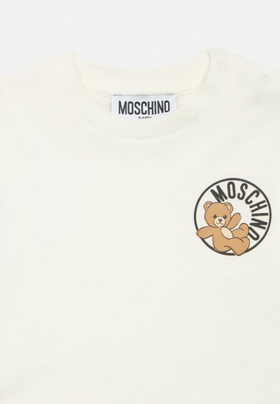 Tricou alb pentru bebeluși, cu nasturi capsa pe umăr, având un ursuleț maro și textul „MOSCHINO” într-un cerc pe piept.