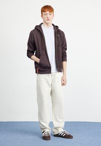 Sudadera con cremallera de color marrón oscuro, con una camiseta blanca debajo, combinada con pantalones cargo de color crema. El modelo está de pie sobre una alfombra azul con un fondo sencillo.