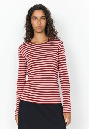 Vrouw met krullend donker haar die een roze en bordeaux gestreept shirt met lange mouwen en een zwarte rok draagt tegen een witte achtergrond.