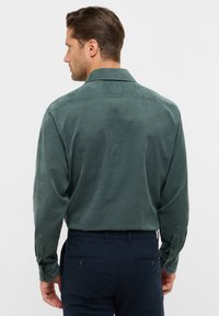 Camicia button-up verde scuro con colletto, maniche lunghe e polsini abbottonati, indossata sopra pantaloni blu navy con un motivo testurizzato e tasche posteriori.