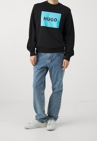 Czarna bluza z niebieskim prostokątnym logo z napisem "HUGO". W zestawie z jasnoniebieskimi dżinsami i białymi sneakersami.