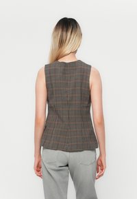 Brun rutete ermeløs vest med en tettsittende design, med rund hals og peplum-lignende silhuett. Komplimentert av grå bukser.