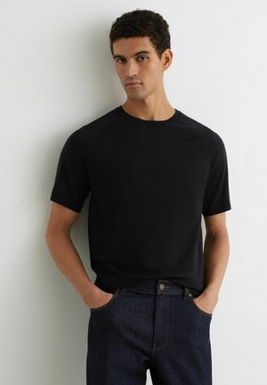 Mann mit lockigem Haar, der ein schwarzes Kurzarmhemd und dunkelblaue Jeans trägt, mit den Händen in den Taschen vor einer weißen Wand stehend.