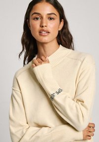Maglione lavorato a maglia color crema con colletto alto a coste e maniche lunghe, caratterizzato da un accento stampato sul polsino e un motivo testurizzato.