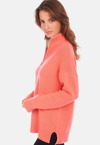 Pull en corail avec un col montant et une demi-fermeture éclair, doté de manches longues, de fentes sur les côtés de l'ourlet, d'une texture côtelée et d'une coupe ample.